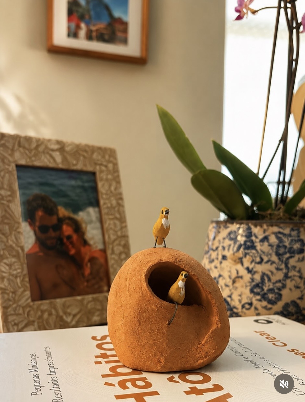 Peça artesanal com passarinhos ao lado de uma fotografia e uma planta